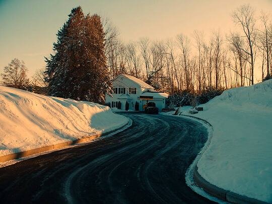 A freshly plowed driveway of bare asphalt curving between deep snow banks toward a white colonial home